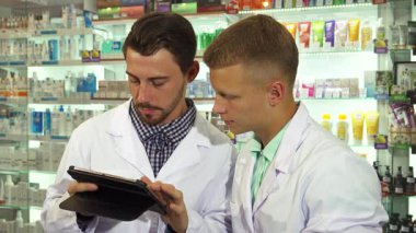 Çıtır beyaz önlüklü iki erkek eczacı, çeşitli ilaçlarla dolu renkli raflarla çevrili bir tableti dikkatle inceliyor. Odaklanmış ifadeleri modern bir ortamda takım çalışması ve profesyonelliği vurguluyor.
