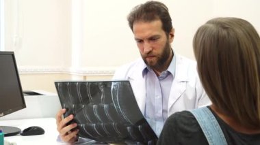 İyi aydınlatılmış bir muayenehanede genç bir kız röntgenini dikkatli bir şekilde ciddi bir terapiste gösterir ve o da bunu dikkatle inceler. Sahne, sağlık danışmanlığı ve bağın önemini vurgulayan bir endişe ve profesyonellik anı yakalıyor.