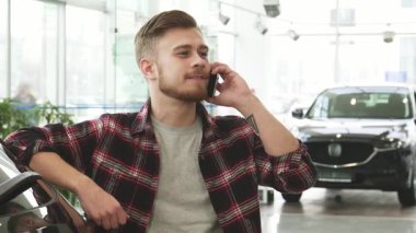 Şık bir ekose gömlek giymiş neşeli, genç sakallı bir adam modern bir araba galerisinde canlı bir telefon görüşmesi yapıyor. Şık araçların arasında heyecan verici haberleri paylaşırken kendine güveni mutluluk saçıyor.