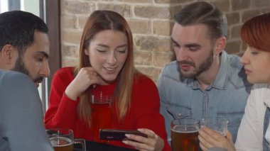 Canlı bir grup genç arkadaş, iki erkek ve iki kadın, rahat bir barda toplanıyor, akıllı telefonlarındaki komik içeriği keşfederken kahkahalarını paylaşıyorlar. Kırsal tuğla duvarlar ve buzlu bira bardaklarıyla vurgulanan ılık ortam mükemmel bir şey yaratıyor.