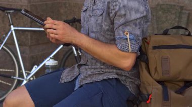 Odaklanmış bir erkek bisikletçi kenarda otururken bir tablet dokunmatik ekranla etkileşime girer, bisikleti yakınlarda park halindedir. Canlı kentsel zemin, teknolojinin ve açık hava yaşam tarzının bir karışımını sergiliyor ve zahmetsizce göz gezdiriyor.