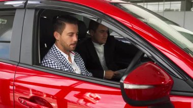 Canlı kırmızı bir arabanın içinde, iyi giyimli bir satıcı ve odaklanmış bir alıcı modern iç mekanı ve çeşitli kontrolleri inceler. Galeri atmosferi arabaların özellikleri hakkındaki tartışmalarını güçlendiriyor, potansiyel bir alışverişin heyecanını vurguluyor.