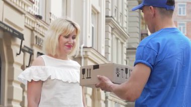 Gülümseyen bir teslimatçıdan dikkatlice paketlenmiş karton bir kutu alan sarı saçlı neşeli bir kadın. Bu canlı açık hava sahnesi online alışverişin heyecanını yakalıyor, müşteri memnuniyeti ile müşteri memnuniyeti arasındaki bağlantıyı gözler önüne seriyor.