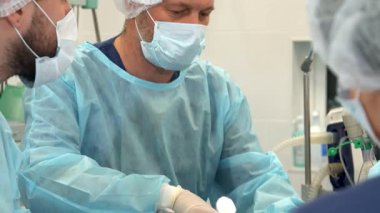 Yüksek riskli bir cerrahi ortamda, kendini adamış bir hemşire cerrahın alnını nazikçe siler, takım çalışmasını gösterir ve odaklanır. Bu yakın plan, steril giyimli ve hayat kurtaran bir prosedüre bağlı iki erkek tıp uzmanını yakalıyor.