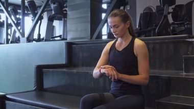 Kararlı genç bir kadın spor salonunun merdivenlerinde güven içinde oturur, saatinin zamanlayıcısını odaklanmış bir ifadeyle ayarlar. Parlak at kuyruğu ve atletik kıyafeti spor yapmaya olan bağlılığını yansıtıyor.