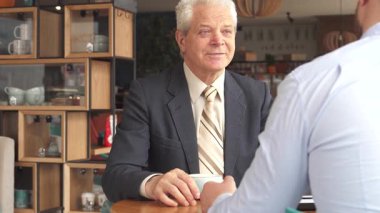 Sıcak bir kafe ortamında, iki Kafkasyalı iş ortağı bir anlık dostluk gösterisi sergileyerek toplantılarını tamamladı. Yaşlı, gri saçlı bir adam masadan kalkan, profesyonelliği somutlaştıran meslektaşıyla tokalaşırken gülümsüyor.