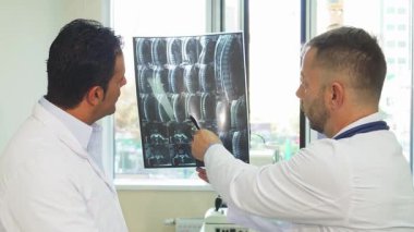 İki deneyimli erkek doktor, iyi aydınlatılmış bir muayenehanede hastalarının sağlıklarına olan bağlılıklarını vurgulamak için röntgen çekiyorlar. Röntgenle ilgili detaylara dikkat çektikleri zaman, olay yeri onların ciddi tartışmalarını yakalıyor.