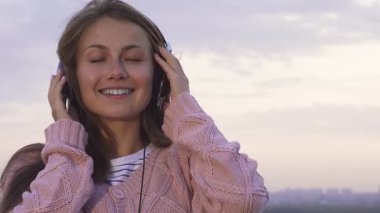 Neşeli genç bir kadın, en sevdiği melodilerle neşeyle dans ederken kulaklıkları yerinde duruyor. Uzun, koyu renk saçları hafif akşam meltemiyle dalgalanır ve çarpıcı şehir silüeti nefes kesici bir zemin görevi görür.