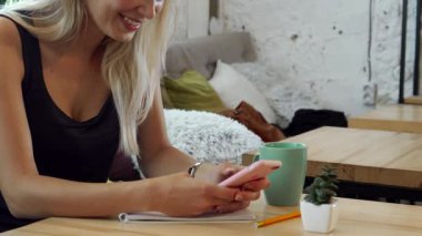 Dalgalı sarı saçlı sakin genç bir kadın rahat bir kafede oturuyor, cep telefonuyla meşgul. Yanında sıcak bir fincan kahveyle, sakinlik ve rahatlama yayıyor, rahatlaması için mükemmel bir ortam yaratıyor.