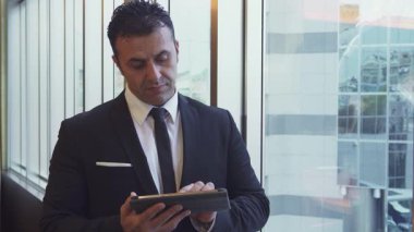 Parlak siyah takım elbiseli ciddi bir adam büyük bir pencerenin yanında durup tabletine yazı yazıyor. Şehir binalarının yansımalarıyla modern ofis ortamı, hırs ve profesyonellik atmosferini artırıyor.