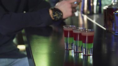 Cilalı bir bar masasında parlayan üç canlı kokteyl fotoğrafının büyüleyici bir yakın çekimi, arka planda oturan bir adam akıllı telefonuyla yazı yazarken. Bu canlı sahne, gece hayatının özünü yakalıyor. Teknoloji ve eğlenceyi modern bir ortamda harmanlıyor.