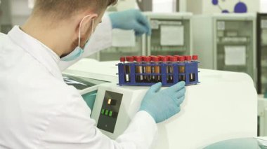 Beyaz önlük ve mavi lateks eldivenler giyen odaklanmış bir laboratuvar teknisyeni, her test tüpünü parlak laboratuvar santrifüjüne yerleştirmeden önce dikkatlice inceler. Bu sahne tıbbi analizde gerekli hassasiyet ve sorumluluğu vurgular.