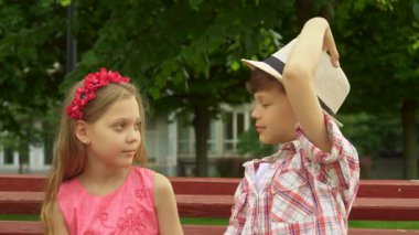 İki neşeli Kafkasyalı çocuk, şık şapkasını ayarlayan yakışıklı bir esmer oğlan ve pembe elbiseli güzel bir kız, canlı kırmızı bir bankta hoş bir gülümseme paylaşıyorlar. Neşeli ifadeleri neşe ve dostluk saçıyor, yemyeşil bir renge karşı.