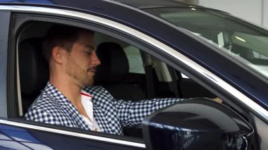 Gösterişli bir galeride sakallı şık bir adam yepyeni arabasında otururken elinde anahtarlarla neşe saçıyor. Kareli gömleği ve parlak gülüşü bu önemli alışverişin heyecanını ve memnuniyetini yansıtıyor.