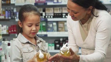 Canlı bir eczanede, genç bir anne ve küçük kızı etiketlerini dikkatlice karşılaştırarak renkli kozmetik tüpleri keşfediyorlar. Paylaştıkları gülücükler ve sohbetleri, güzellik ürünleri seçmenin verdiği keyfi vurgulayan sıcak bir atmosfer yaratıyor.