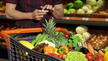 Neşeli bir gülümseme ile şık bir genç adam, canlı meyve ve sebze dolu canlı bir market arabasının yanında dururken akıllı telefonuyla alışveriş listesini kontrol eder. Özenli bakışları ve günlük tavırları alışverişin heyecanını vurguluyor.