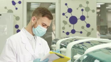 Çıtır beyaz önlük ve mavi lateks eldivenler giyen genç erkek laboratuvar asistanı gelişmiş laboratuvar makinelerinin yanında duruyor. Test sonuçlarını dikkatlice parlak sarı bir klasöre kaydediyor. Hassasiyet ve adanmışlığı somutlaştırıyor.