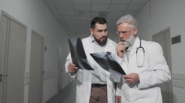 Parlak bir hastane koridorunda genç bir erkek doktor ve deneyimli kıdemli meslektaşı, x-ray taramalarını kasıtlı ifadelerle analiz ederek sohbete daldılar. Beyaz önlükleri ve steteskopları profesyonelliği simgeler.
