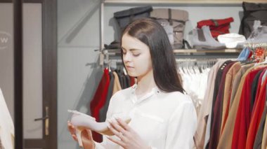 Uzun siyah saçlı zarif bir kadın şık bir butikte şık bir yüksek topuklu ayakkabıdan etkilenir. Canlı kıyafetler ve aksesuarlarla çevrelenmiş, modern moda ve kişisel tarzın özünü somutlaştırıyor.