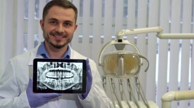 İyi bakımlı sakallı neşeli orta yaşlı bir beyaz dişçi modern bir klinikte temiz bir diş röntgeni gösteren bir tablet tutuyor. Parlak ofis, diş teçhizatlarıyla donatılmış, davetkar atmosferi arttırıyor,