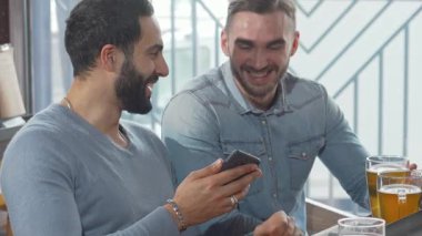 İki neşeli erkek arkadaş şık bir barda oturmuş soğuk biralarını yudumluyor ve bir akıllı telefona dalmış durumdalar. Kahkahaları ve yoldaşlıkları, arkadaşlığın ve modern iletişimin neşesini gösteren canlı bir atmosfer yaratıyor.