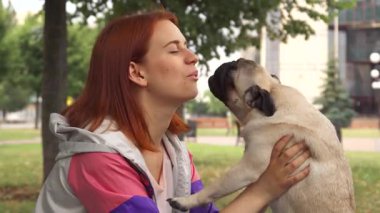 Neşeli bir kız, sevgili köpeğini yüzüne yakın bir yere yatırır. Neşeli yavru, dudaklarını yalamaya çalışırken neşeyle ışıldar. Bu yürek ısıtan an, güneşli bir parkta yemyeşil bir alanla çevrelenmiş bağlarının özünü yakalar.