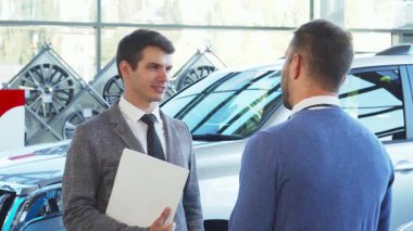 Parlak araçlarla dolu şık bir araba galerisinde, iyi giyimli bir satıcı bir alıcıyla canlı bir tartışmaya girer. Satıcının anahtarları teslim ettiği an, başarılı bir anlaşmayı ve yeni bir heyecanı simgeliyor.