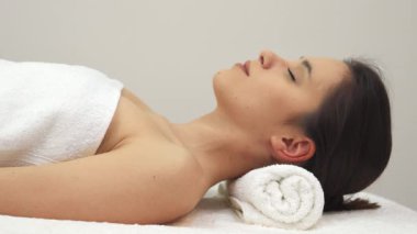 Çekici esmer bir kız, yumuşak beyaz bir havluya sarılı masaj masasında uzanmış, ışıl ışıl gülüşü, huzurlu spa atmosferini aydınlatıyor. Bu çekici beyaz kadın model yüzünü kameraya çeviriyor, rahatlama ve güzelliği somutlaştırıyor.