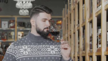 Şık sakallı bir adam şık şişelerle dolu raflarla çevrili, hareketli bir şarap süpermarketinde bir bardak zengin kırmızı şarabın tadına zarif bir şekilde bakar. Kendine güveni ve rahat süveteri güzel şaraplara olan tutkusunu yansıtıyor.