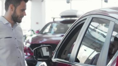 Modern bir galeride şık bir arabaya yaslanmış çekici bir adam kameraya ışıldıyor. Neşeli tavrı yeni bir araç seçmenin heyecanını yansıtıyor, araba sahibi olmanın heyecanını simgeliyor.