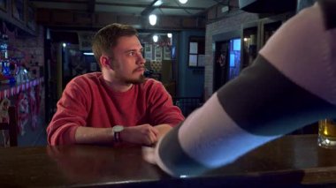 Bir barmen eli titizlikle parlayan bir yüzey oluşturarak parlayan tezgahı siler. Arka planda, rahat kırmızı kazaklı genç beyaz bir adam düşünceli bir bira yudumlar, bir spor oyununun heyecanına kapılır,