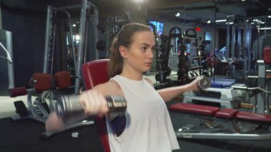 Uzun sarı saçlı, at kuyruklu kararlı genç kadın, yoğun bir şekilde dambılları kaldırıyor, gücünü ve bağlılığını gösteriyor. Spor malzemeleriyle çevrili, spor ruhunun vücut bulmuş hâli, her temsilciden sınırlarını zorluyor.