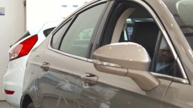 Zarif sakallı ve gözlüklü çekici bir beyaz adam parlak gri bir sedanı incelemek için eğildi ve onayladı. Kendine güveni ve profesyonel kıyafeti modern galeride davetkar bir atmosfer yaratıyor.