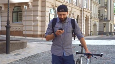 Yakışıklı sakallı bir bisikletçi resimli bir şehir sokağında cep telefonunu dikkatle kontrol ediyor. Bu çekici genç adam, şık bir şapka ve sırt çantasıyla şık bisikletinin yanında duruyor, fitness ve modern yaşam tarzının bir karışımını somutlaştırıyor.