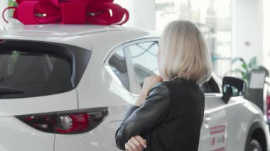Işıl ışıl gülümseyen kendine güvenen bir kadın galeride canlı bir fiyonkla süslenmiş şık ve yeni bir arabayı inceliyor. Neşeli tavrı, mükemmel arabayı seçmenin heyecanını yansıtıyor. Tarz ve entelektüelliğin bir karışımı.