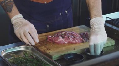 Kesilmiş bir üst manzara, mavi önlüklü yetenekli bir şefi ahşap kesme tahtasına taze domuz eti serpiştirirken ve aromatik biberiyeyle çevrili bir şekilde yakalıyor. Bu aşçılık anı lezzet hazırlığının sanatsallığını vurguluyor.