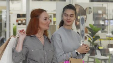 Biri canlı kızıl saçlı, diğeri şık gri elbiseli iki neşeli genç kadın, alışveriş merkezinde sergilenen şık eşyalardan büyüleniyor. Gülüşleri ve gülüşleri alışverişin heyecanını simgelerken heyecanları yayılıyor.