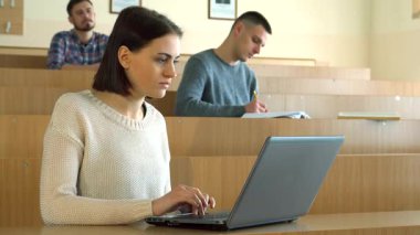 Çekici beyaz bir kadın, arka planda iki erkek sınıf arkadaşı çalışırken, dizüstü bilgisayarıyla derinden ilgilenen ahşap bir masada oturuyor. Parlak sınıf ayarları konsantrasyon ve akademik takip atmosferini yükseltiyor.