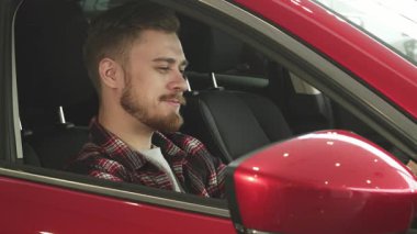 Düzgün bakımlı sakallı şık bir genç adam canlı kırmızı arabasının sürücü koltuğunda rahatça oturur, neşe ve memnuniyet saçar. Rahatlamış tavrı ve büyüleyici gülüşü yeni sahipliğin heyecanını yansıtıyor.