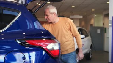 Neşeli, kıdemli, beyaz bir adam galeride canlı, mor bir kavşağın arka kapağını sevinçle kaldırıyor. Onun parlak gülümsemesi modern bir sergi salonu atmosferiyle çevrili geniş bagaj kompartımanını keşfetme heyecanını yansıtıyor.