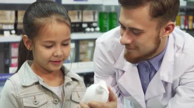 Neşeli küçük bir kız gülümseyen bir eczacının yanında durur ve bir şişe sıvı ilacı birlikte inceler. Çekici sohbetleri sıcaklık ve merak yayıyor, renkli ürünlerle dolu canlı eczanede keyifli bir an yaratıyor.
