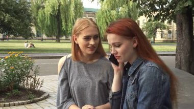 Kızıl saçlı bir kız akıllı bir telefonla sohbet etmek için öne eğilirken, sarışın arkadaşı yanında oturmuş, canlı bir tartışmayla meşgul oluyor. Üniversite parkında yemyeşil bir ortamda, hareketli ifadeleri arkadaşlığı ve neşeyi yansıtıyor.