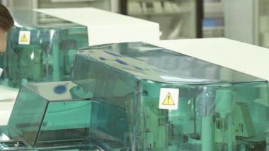 Maske ve lateks eldiven takan kendini işine adamış genç bir erkek laboratuvar asistanı dönen santrifüje yakından bakıyor. Ekipmanların canlı yeşil kaplaması içerideki temiz borularla çelişiyor.