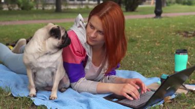 Neşeli bir kız güneşli bir parkta canlı mavi bir battaniyenin üzerinde rahatlar, sevimli köpeği ona bakarken dikkatle dizüstü bilgisayarında yazar. Sahne sıcaklığı ve arkadaşlığı yansıtıyor. Doğada verimliliğin ve eğlencenin mükemmel bir karışımını sergiliyor.