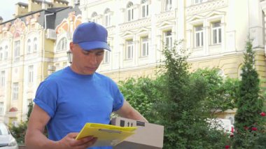 Canlı mavi gömlekli ve şapkalı odaklanmış bir erkek kurye elinde bir karton kutuyla paketi teslim etmeye hazırlanırken çevreyi tarıyor. Şehir arkaplanı büyüleyici bir mimari ve yemyeşil bir ortam içeriyor, bu hareketli olaya canlı bir dokunuş katıyor.