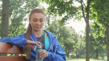 Işıl ışıl bir sporcu kadın, su şişesinden ferahlatıcı bir yudum alır ve enerji dolu bir hava egzersizinden sonra güneşin altında güneşlenir. Canlı mavi spor kıyafetleri etrafındaki yemyeşil yeşilliği tamamlıyor. Sağlığını, canlılığını ve kalmanın verdiği keyfi temsil ediyor.