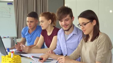 Şık bir ofis çalışma alanında canlı bir işbirliği içinde olan genç profesyonellerin canlı bir sahnesi. Mavi tişörtlü yakışıklı esmer bir adam dizüstü bilgisayardaki konseptleri odaklanmış kızıl bir kadına anlatırken, sakallı çekici bir adam...