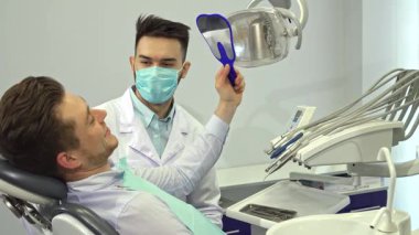 Aynadaki yansımasında sarı saçlı yakışıklı genç bir adam dişçi ofisinde parlak gülümsemesini sergiliyor. Maske takıp önlük giyen, özenli esmer, beyaz dişçi, dişçi aletleri ile hazır bekliyor.