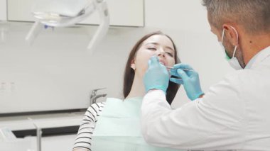 Neşeli genç bir kadın dişçi koltuğunda rahatlıyor, parlak gülüşü dişlerini inceleyen yetenekli bir erkek dişçi gibi kendine güven saçıyor. Modern klinik çevresi, parlak ekipmanları ve sakinleştirici renkleriyle,