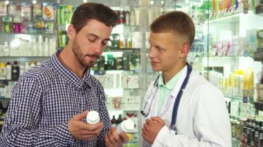 Parlak bir eczanede, beyaz önlüklü bir erkek doktor iki hap şişesini hastasıyla tartışıyor, o da bunları dikkatle karşılaştırıyor. Atmosfer profesyonel ama dost canlısı, sağlık hizmetlerinde bilinçli seçimlerin önemini vurguluyor.